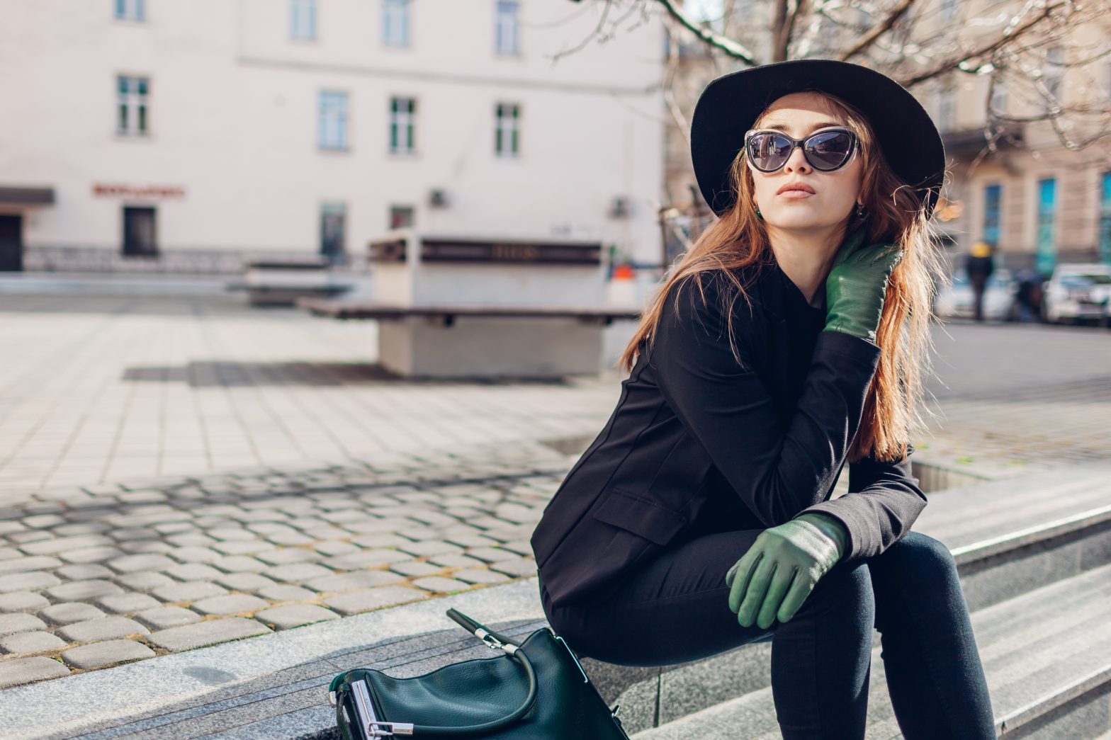 Young woman in black clothes, sunglasses and hat, wearing green gloves, sits in an European town square.